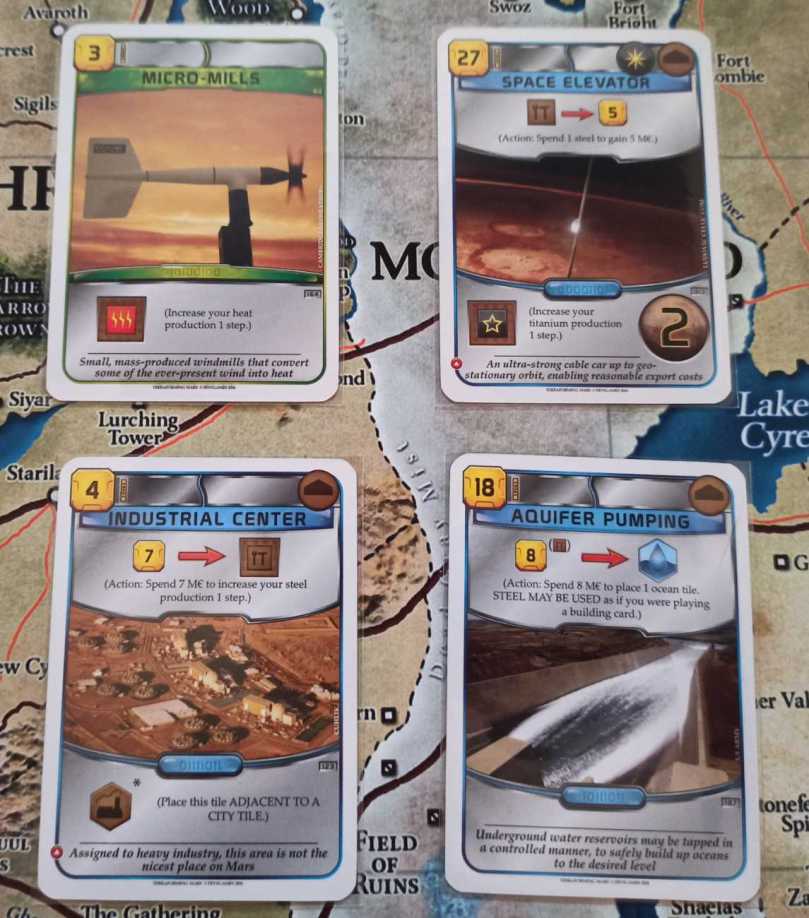 From top left moving clockwise, the cards are: Micro Mills, costing 3 credits and giving 1 heat production. Space Elevator, costing 27 credits, giving 2 VP and 1 titanium production, and an action that allows to spend 1 steel for 5 credits. Industrial Center, costing 4 credits and placing a special tile next to a City, and gives an action that increases your steel production by 1 for 7 credits. Aquifer Pumping, costing 18 credits and giving an action that places an Ocean tile for 8 credits, allowing to pay with steel as well.